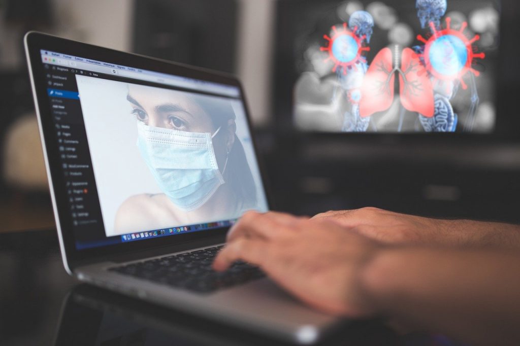 Zdjęcie nawiązujące do epidemii koronawirusa – komputer z wizerunkiem kobiety w masce oraz w tle obraz przedstawiający płuca i wirusy