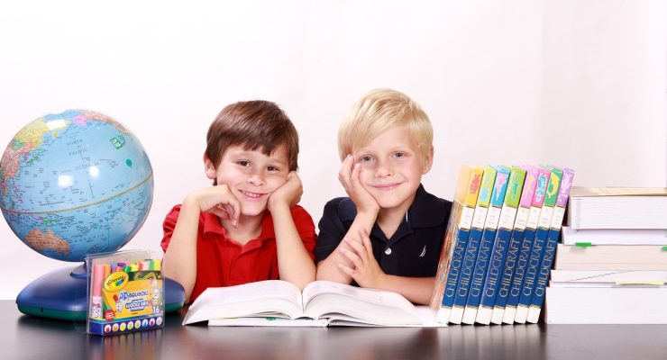 Zdjęcie przedstawia dwóch chłopców siedzących przy biurku i przeglądających książkę. Na biurku znajdują się: globus, książki i flamastry.
