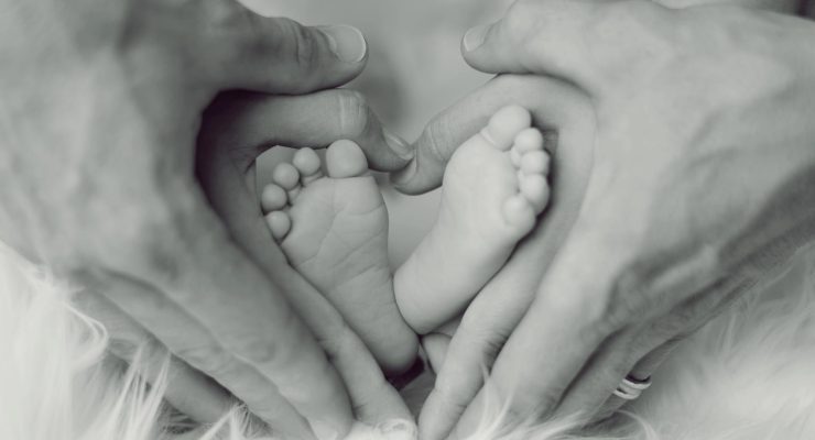 Grafika przedstawiająca ręce kobiety i mężczyzny obejmujące stópki niemowlaka.