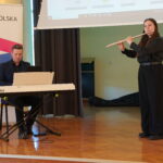 Występ muzyczny – mężczyzna gra na keyboardzie a młoda kobieta gra na flecie poprzecznym. Oboje ubrani na czarno, stoją na scenie w sali konferencyjnej z banerami promującymi Małopolskę w tle. Za nimi znajduje się ekran projekcyjny